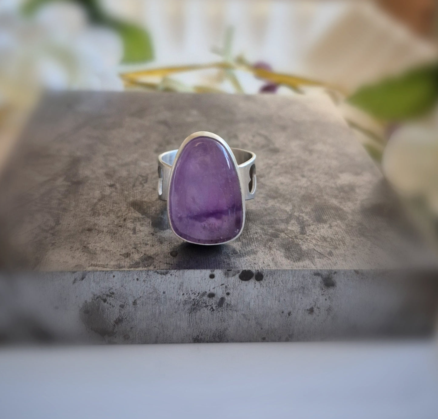 Silver ring with a purple amethyst stone on a gray surface with flowers in the background