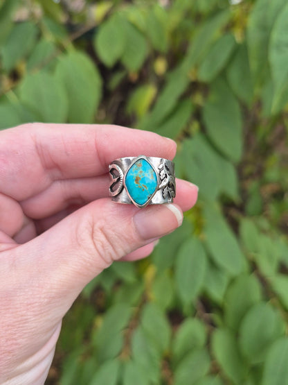 Hand holding a silver ring with turquoise stone against a green leafy background