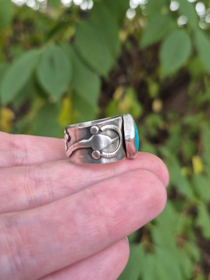 Silver ring with turquoise stone held between fingers against a blurred green leafy background