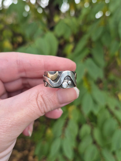 Hand holding a silver ring with a wavy design against a blurred green leafy background