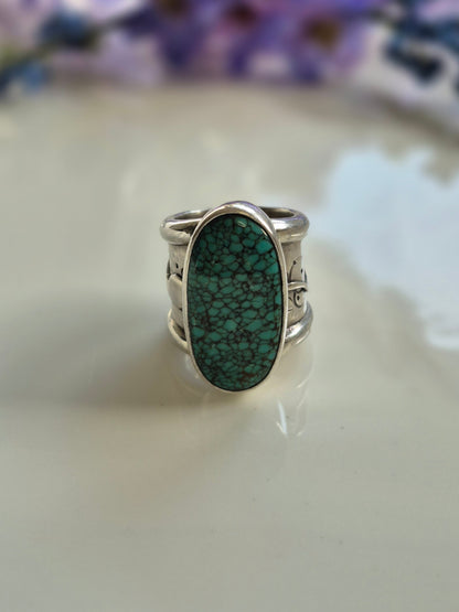 Silver ring with a green turquoise stone on a white surface with a blurred purple flower in the background