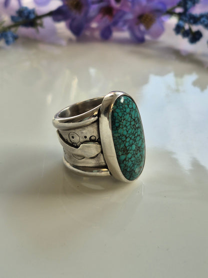 Silver ring with a green turquoise stone on a white surface with purple flowers.