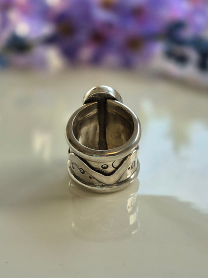 Silver ring with intricate designs on a white surface with a blurred purple flower background