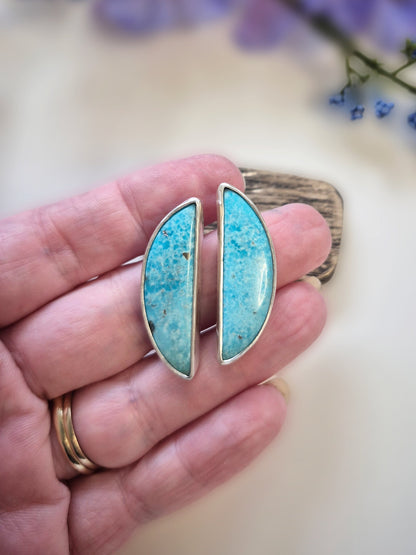 Turquoise earrings held in a hand with a blurred floral background