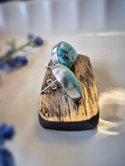 Turquoise stone earring on a wooden stand with blurred blue flowers in the background