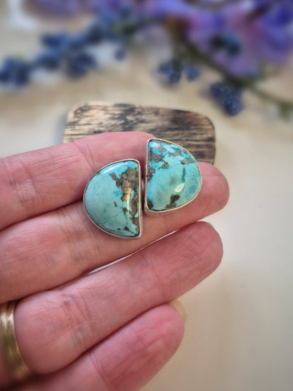 Turquoise stone earrings held between fingers with a blurred floral background