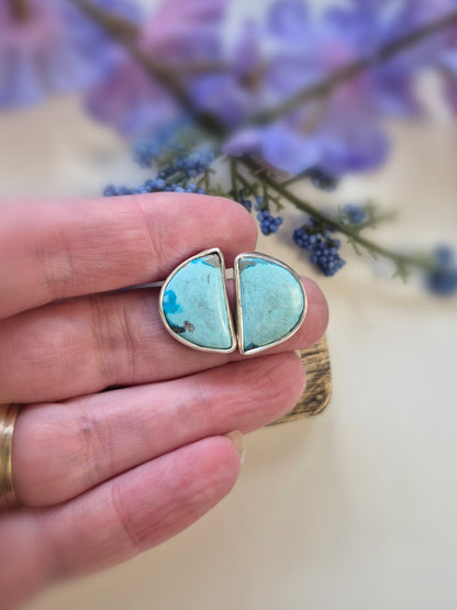 Turquoise half-circle earrings held between fingers with a blurred floral background