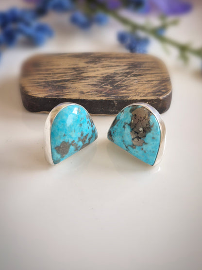 Turquoise gemstone earrings with silver settings on a white surface with blurred blue flowers in the background.