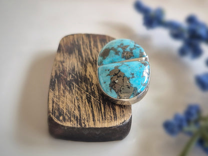 Turquoise stone ring on a wooden stand with blurred blue flowers in the background