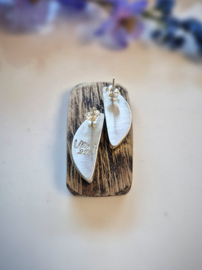 Silver crescent-shaped earrings on a wooden stand with blurred purple flowers in the background