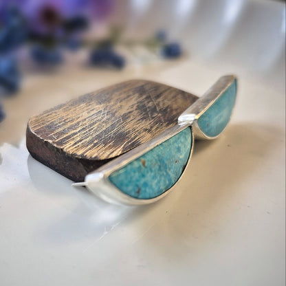 Earrings and turquoise gemstones against a wooden platform on a reflective surface with blurred flowers in the background