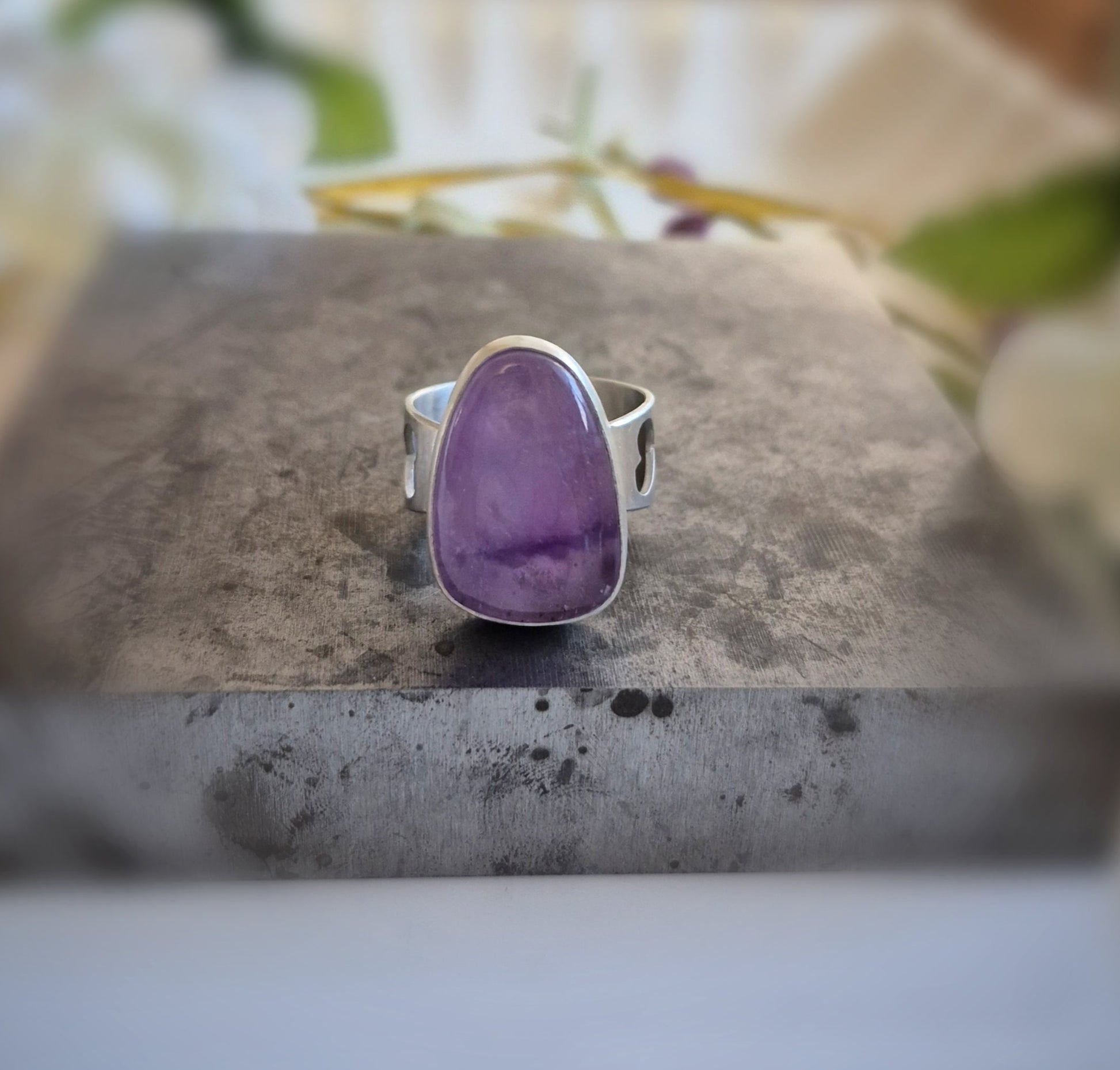 Silver ring with a purple amethyst stone on a gray surface with flowers in the background