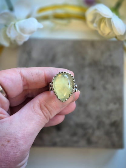 Hand holding a ring with a yellow prehnite gemstone against a blurred background