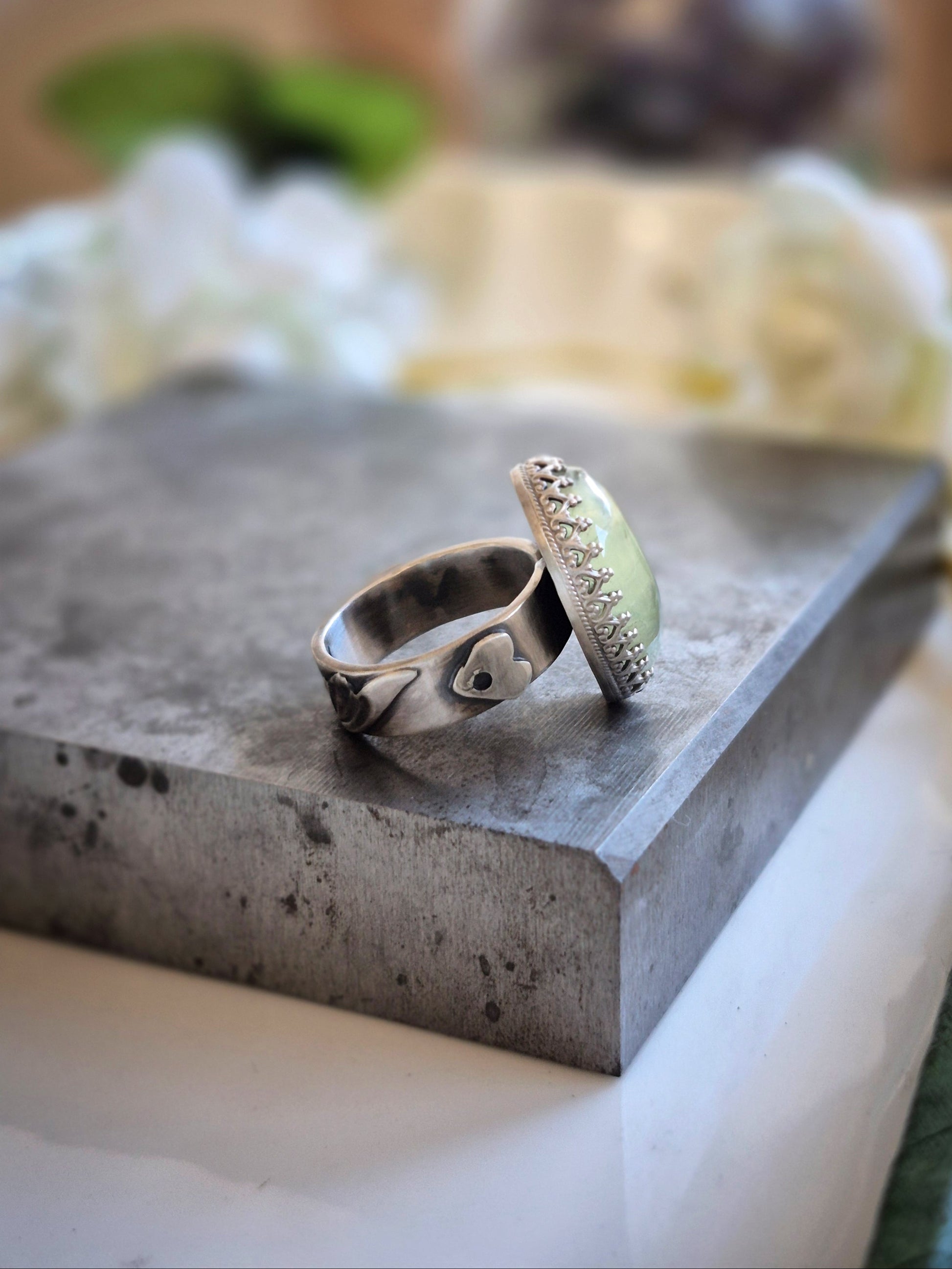 A silver ring with a heart cutout on side of band and a fancy bezel on a concrete surface with a blurred natural background
