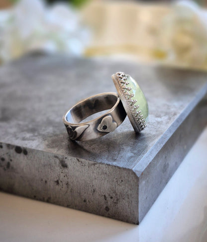 A silver ring with a heart cutout on side of band and a fancy bezel on a concrete surface with a blurred natural background